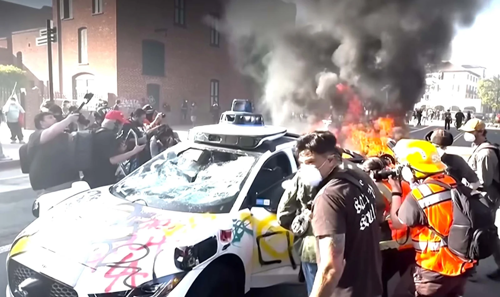 A damaged autonomous car surrounded by smoke and a crowd